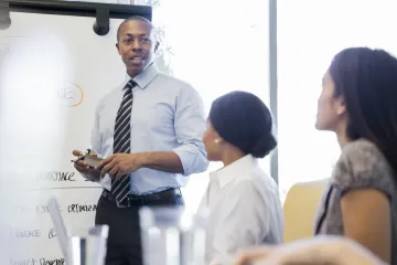 Person standing at whiteboard speaking with two seated people at work.